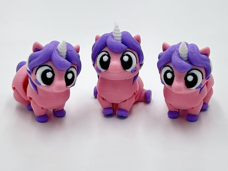 Peut inclure: Trois petites figurines de licorne imprim&eacute;es en 3D avec des corps roses, des crini&egrave;res violettes et des cornes blanches. Chaque licorne a de grands yeux noirs et blancs, de petits sabots violets et une posture assise.