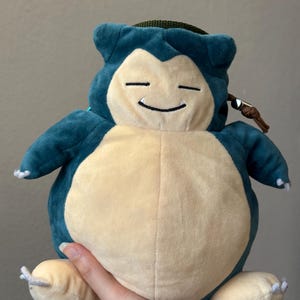 May include: A Snorlax plush toy with a teal head and limbs, a cream-coloured belly, and a green top. The toy has a smiling face with closed eyes and a drawstring closure. The toy is held by a person's hand.