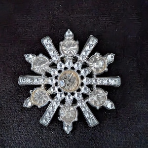 May include: A silver-toned brooch shaped like a snowflake, featuring a central clear round rhinestone surrounded by six smaller clear round rhinestones. The metal frame is embellished with numerous tiny clear crystals along the radiating arms.