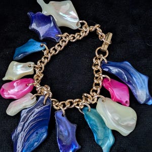 May include: A gold-toned chain bracelet adorned with colorful, iridescent shell-shaped charms. The charms are in shades of blue, pink, and white, creating a vibrant and eye-catching accessory. The bracelet has a lobster clasp closure.