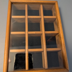 May include: A rectangular wooden display box featuring a clear glass lid and a black interior lining. The interior is divided into twelve equal compartments by a grid of wooden dividers, providing organized storage space for small items.