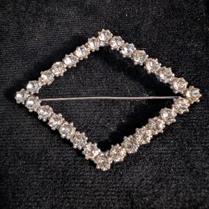May include: A diamond-shaped brooch featuring a silver-toned metal frame set with numerous clear, faceted rhinestones. The piece includes a horizontal pin mechanism spanning the center of the open geometric frame, showcasing a classic design.