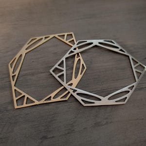 May include: Two geometric metal frames, one in a gold tone and one in a silver tone. Each frame features an openwork design with a hexagonal outer shape and intricate, angular cutouts forming a complex pattern within the border structure.
