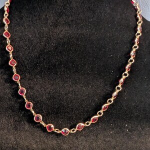 May include: A gold-toned chain necklace featuring a series of evenly spaced, round, deep red faceted stones set in metal bezels. The necklace is constructed with simple circular links connecting each individual stone setting in a continuous loop.
