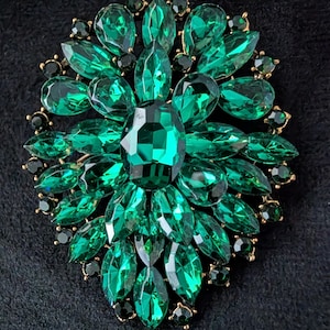 May include: A circular brooch featuring a large central emerald green faceted crystal surrounded by multiple layers of marquise and pear-shaped green crystals. The stones are set in a gold-toned metal frame with small dark green accent crystals.