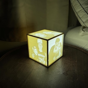 May include: A glowing, square lamp with multiple black and white photographs displayed on its sides. The lamp is illuminated with a warm, yellow light, showcasing the images. The lamp sits on a dark wooden surface.