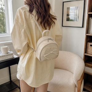 May include: A cream-coloured mini backpack featuring a heart-shaped design and bow detail. The small backpack has a top handle, a zipped closure, and adjustable straps. The heart design is embellished with lace trim.