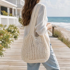 May include: Cream-coloured, open-weave crochet tote bag with a long shoulder strap. The bag is carried over the shoulder. The bag's design features a grid-like pattern. The background shows a beach scene with a wooden walkway and ocean.