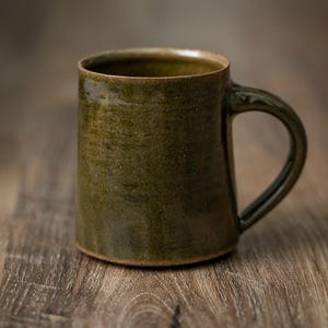 May include: A cylindrical ceramic mug with a dark olive green glaze and a glossy finish. The mug features a sturdy, curved handle on the right side and a slightly textured, earthy surface with subtle horizontal markings throughout the exterior body.