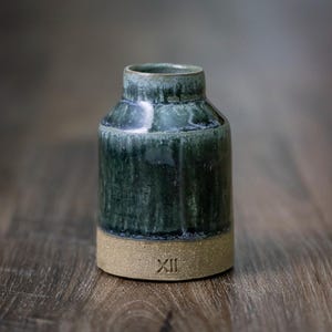 May include: A handcrafted ceramic vase with a dark green glaze over a tan base. The vase has a cylindrical shape with a slightly flared rim. The base has the Roman numeral XII. The vase is approximately 5 inches tall.