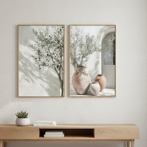 May include: Two framed art prints with gold frames hang above a wooden media console. The prints depict olive trees and terracotta vases. A small potted plant and a beige rug are in the foreground. The room has white walls and a white floor.
