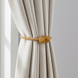 May include: A pair of light beige curtains held together by a gold-colored rope tieback with geometric wooden accents. The curtains are hanging in front of a white window frame.