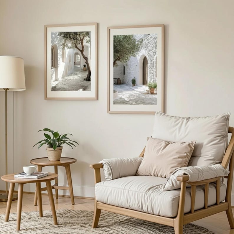 May include: Two framed wall art prints depicting Mediterranean stone courtyards with olive trees, displayed above a light wood armchair with beige cushions, two round wooden side tables, and a floor lamp with a cream-colored shade.