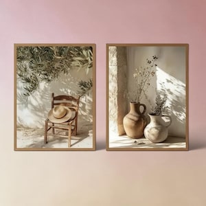 May include: A set of two framed wall art prints. The left print shows a wooden chair with a straw hat resting on the seat under olive tree branches. The right print displays two ceramic vases containing dried stems against a stone wall.