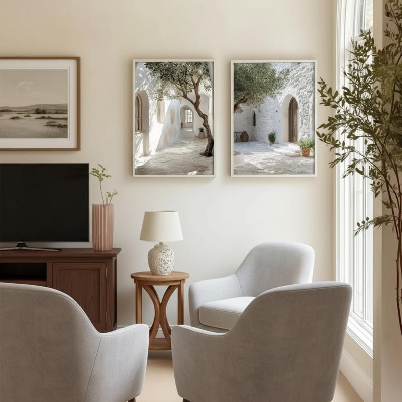 May include: A living room featuring two light gray armchairs, a wooden media console with a television, a small round wooden side table with a white lamp, and two framed wall art prints depicting Mediterranean stone courtyards and olive trees.