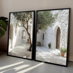 May include: Two rectangular wall art prints in black frames featuring a Mediterranean scene with white stone buildings, arched doorways, a large olive tree, and a paved courtyard. The imagery displays bright sunlight casting shadows on the stone ground.