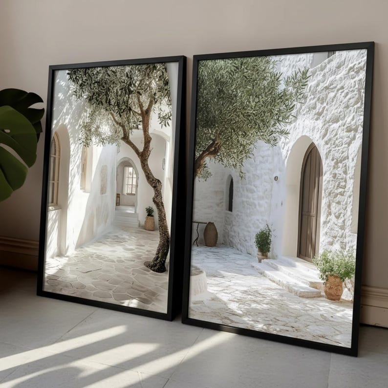 May include: Two rectangular wall art prints in black frames featuring a Mediterranean scene with white stone buildings, arched doorways, a large olive tree, and a paved courtyard. The imagery displays bright sunlight casting shadows on the stone ground.
