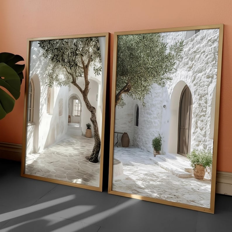 May include: Two rectangular framed wall art prints featuring a Mediterranean courtyard scene with white stone walls, a gnarled olive tree, a dark wooden arched door, and a paved stone ground. The frames are gold-toned and lean against a peach wall.