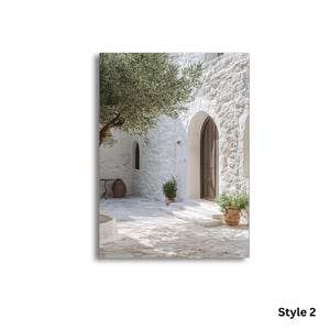 May include: A white stone building with an arched wooden doorway and stone patio. A large olive tree with green leaves hangs over the courtyard. Two potted green plants sit near the entrance, and a dark brown vase stands in the corner.