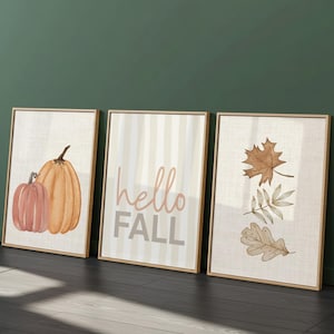 May include: Three framed wall art prints with light wood frames. The left print shows two orange pumpkins. The centre print features vertical stripes and the text hello FALL in brown. The right print displays three brown autumn leaves.