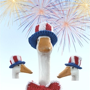 May include: Three white goose figurines with orange beaks, each wearing a crocheted top hat with red and white vertical stripes and a blue brim. The central goose also wears a matching red and white crocheted bow tie against a sky background.