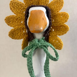 May include: A crochet sunflower bonnet worn by a white goose figurine. The bonnet features bright yellow petals, a dark brown center frame, and a light green crocheted tie that is knotted securely under the chin of the goose.