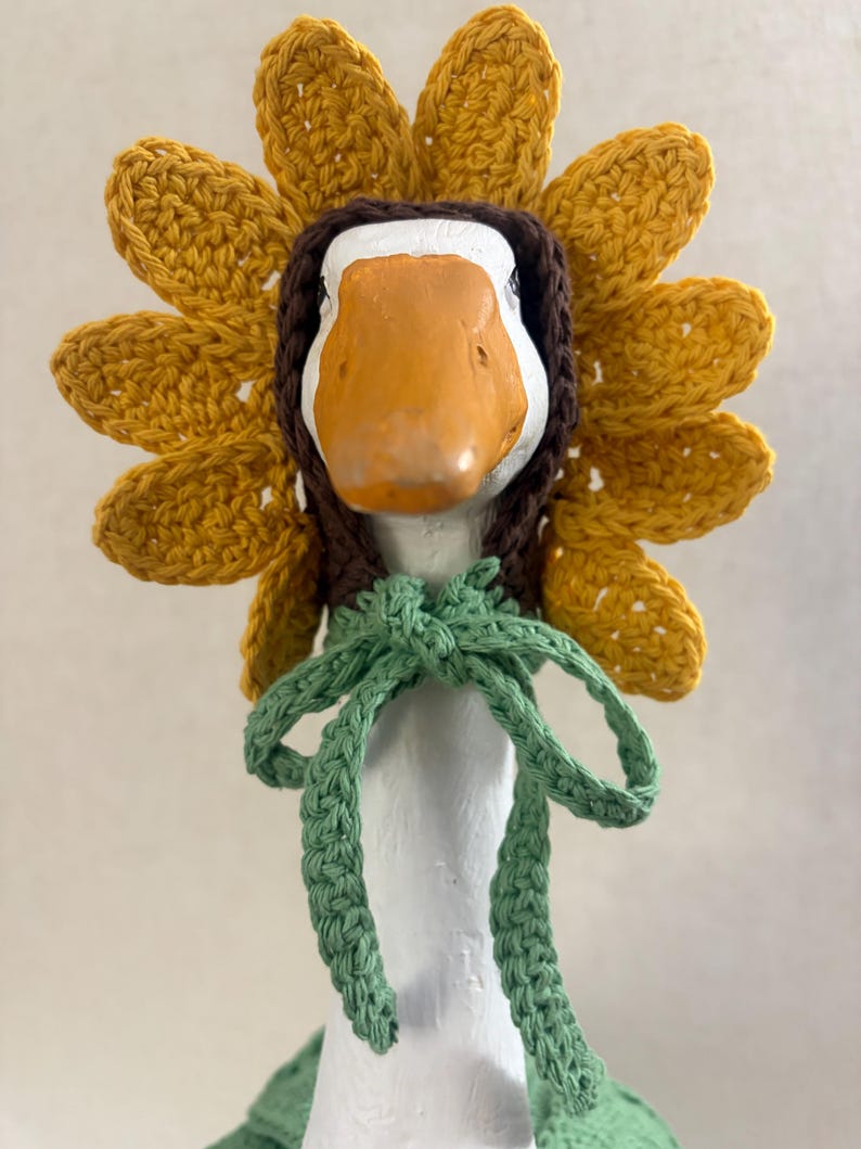 May include: A crochet sunflower bonnet worn by a white goose figurine. The bonnet features bright yellow petals, a dark brown center frame, and a light green crocheted tie that is knotted securely under the chin of the goose.
