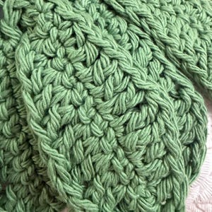 May include: A sage green crocheted textile featuring a dense, repeating stitch pattern. The yarn is thick and creates a textured, looped surface with visible interconnected strands forming a consistent, rhythmic design across the entire fabric.