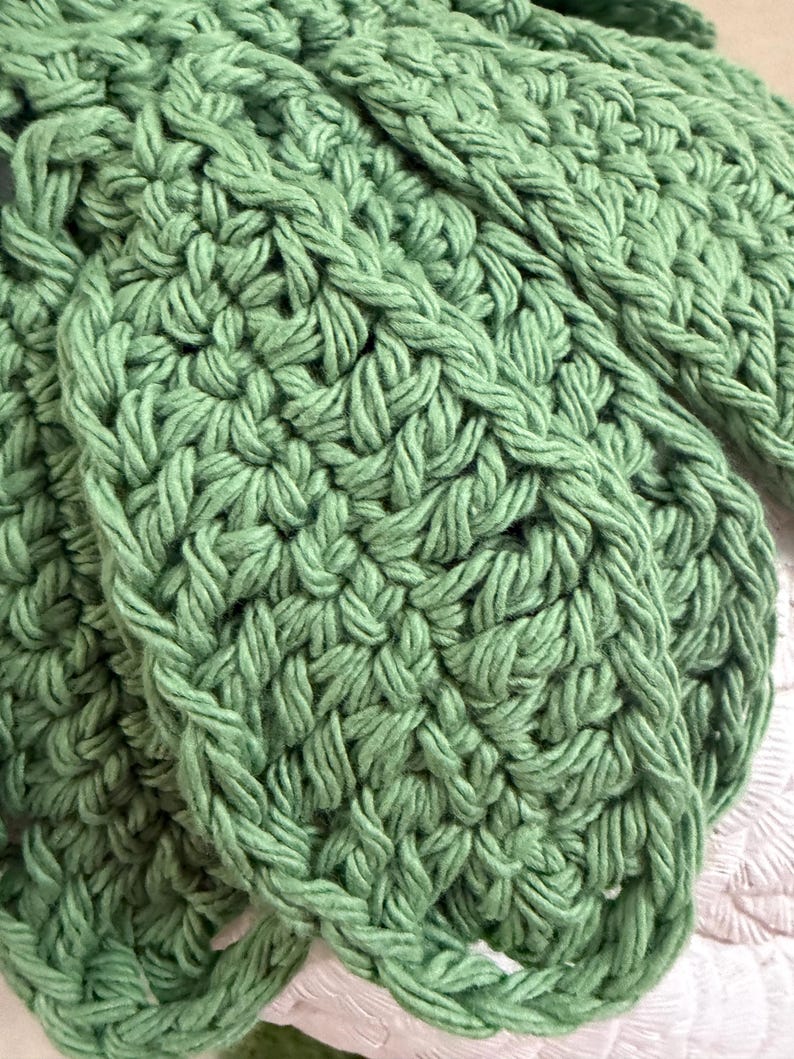 May include: A sage green crocheted textile featuring a dense, repeating stitch pattern. The yarn is thick and creates a textured, looped surface with visible interconnected strands forming a consistent, rhythmic design across the entire fabric.
