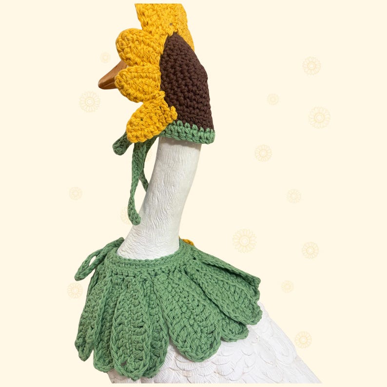 May include: A white goose figurine wearing a crocheted sunflower hat with yellow petals and a brown center, paired with a green crocheted leaf collar. The hat features long green ties, and the entire set is crafted from textured yarn.