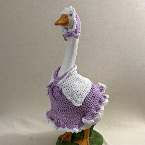 May include: A white goose figurine wearing a handmade crochet outfit. The outfit includes a lavender dress with white trim, a matching bonnet, and a white bib. The goose has an orange beak and feet, and the base is green.