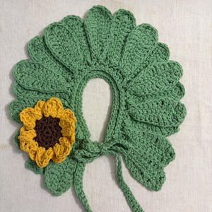 May include: A crochet collar featuring a series of sage green leaf-shaped petals arranged in a circular pattern. A single three-dimensional sunflower with yellow petals and a dark brown center is attached to the left side. Two thin ties hang down.