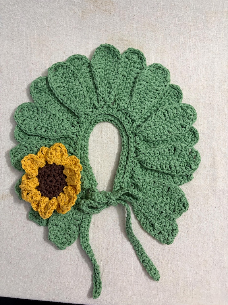 May include: A crochet collar featuring a series of sage green leaf-shaped petals arranged in a circular pattern. A single three-dimensional sunflower with yellow petals and a dark brown center is attached to the left side. Two thin ties hang down.