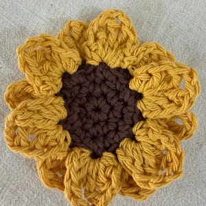 May include: A handcrafted crochet sunflower coaster featuring a dark brown circular center and vibrant yellow petals arranged in a layered, circular pattern. The yarn texture is visible throughout the piece, showcasing the intricate stitch work.