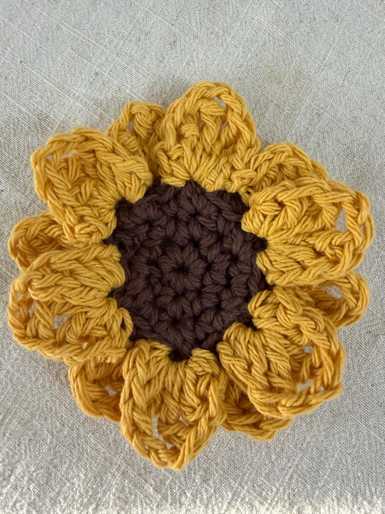 May include: A handcrafted crochet sunflower coaster featuring a dark brown circular center and vibrant yellow petals arranged in a layered, circular pattern. The yarn texture is visible throughout the piece, showcasing the intricate stitch work.