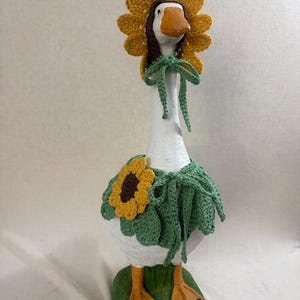 May include: A white goose figurine wearing a crocheted sunflower bonnet with a brown center and yellow petals, tied with green strings. The goose also wears a green crocheted capelet featuring a large sunflower appliqu&eacute; and green ties.