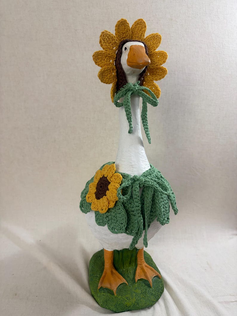 May include: A white goose figurine wearing a crocheted sunflower bonnet with a brown center and yellow petals, tied with green strings. The goose also wears a green crocheted capelet featuring a large sunflower appliqu&eacute; and green ties.
