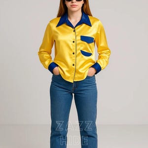 May include: A yellow satin shirt with a blue collar, cuffs, and pocket detail. The shirt has black buttons and is paired with blue jeans. The text "ZAZZ HIDE" is visible on the jeans.