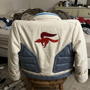 Pode incluir: Um casaco de cor creme com um emblema de raposa vermelho bordado nas costas. A peça inclui painéis acolchoados azuis na parte inferior e um detalhe de risca azul na gola. O design é estruturado com mangas compridas e punhos.