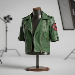 May include: A short-sleeved green leather jacket featuring a notched lapel, gold-toned metal zippers, and a circular patch on the left sleeve with a red and black X design. The jacket is displayed on a dark brown mannequin torso stand.