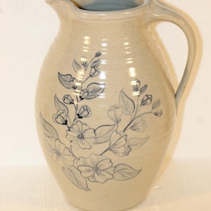 May include: A beige ceramic jug with a handle and spout. The jug is decorated with a blue floral design. The jug is likely used for serving beverages and has a rustic, handmade appearance.