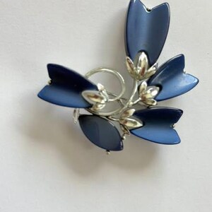 Könnte beinhalten: Eine florale Brosche mit fünf glatten, dunkelblauen, tulpenförmigen Blütenblättern an einem silberfarbenen Metallstiel. Das Design enthält kleine silberfarbene Metallknospen, die zwischen den blauen Blütenblättern sitzen.