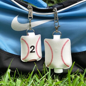 May include: Two white baseball-themed hand sanitizer holders with red stitching and black numbers. The holder on the left has the number "2" on it. Both holders are attached to silver keychains and are hanging from a blue and white sports bag.