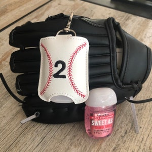 May include: A white baseball-themed hand sanitizer holder with the number "2" embroidered on it. The holder is attached to a black baseball glove and a small bottle of pink PocketBac hand sanitizer that says "Sweet as Strawberries" is in front of the glove.