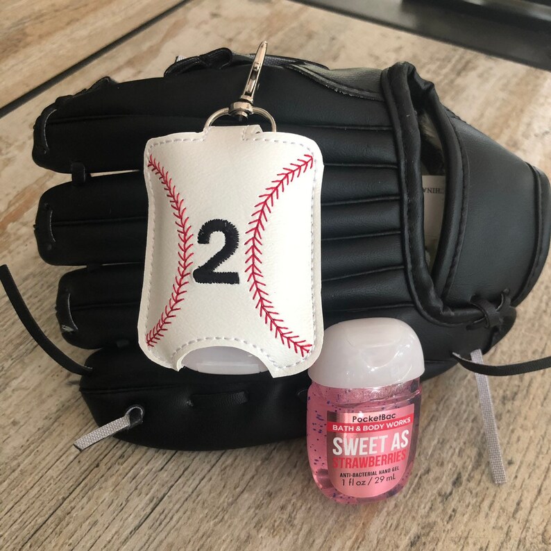 May include: A white baseball-themed hand sanitizer holder with the number "2" embroidered on it. The holder is attached to a black baseball glove and a small bottle of pink PocketBac hand sanitizer that says "Sweet as Strawberries" is in front of the glove.