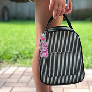 Puede incluir: Una bolsa de almuerzo a rayas blancas y negras con cierre de cremallera y una etiqueta de nombre rosa y blanca que dice "ZOE".