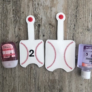 May include: Two white baseball-themed hand sanitizer holders with red stitching and the number "2" on one holder. The holders are on a wood background. A pink hand sanitizer bottle and a clear hand sanitizer bottle are also shown.