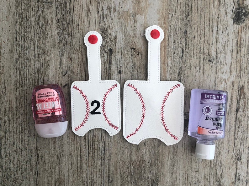 May include: Two white baseball-themed hand sanitizer holders with red stitching and the number "2" on one holder. The holders are on a wood background. A pink hand sanitizer bottle and a clear hand sanitizer bottle are also shown.
