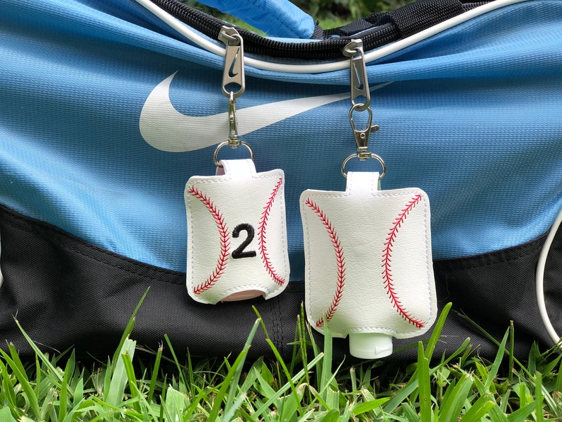 May include: Two white baseball-themed hand sanitizer holders with red stitching and black numbers. The holder on the left has the number "2" on it. Both holders are attached to silver keychains and are hanging from a blue and white sports bag.