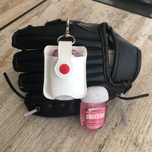 May include: A white leather hand sanitizer holder with a red button attached to a black baseball glove. A pink bottle of PocketBac Sweet as Strawberries anti-bacterial hand gel is next to the holder. The bottle is 1 fl oz / 29 ml.